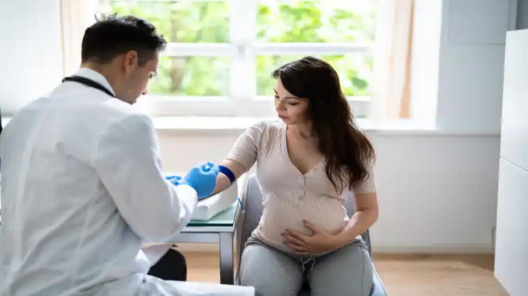 Auf dem Bild sitzt eine junge schwangere Frau auf einem Sessel neben einem Arzt, der ihr gerade Blut abnehmen möchte.